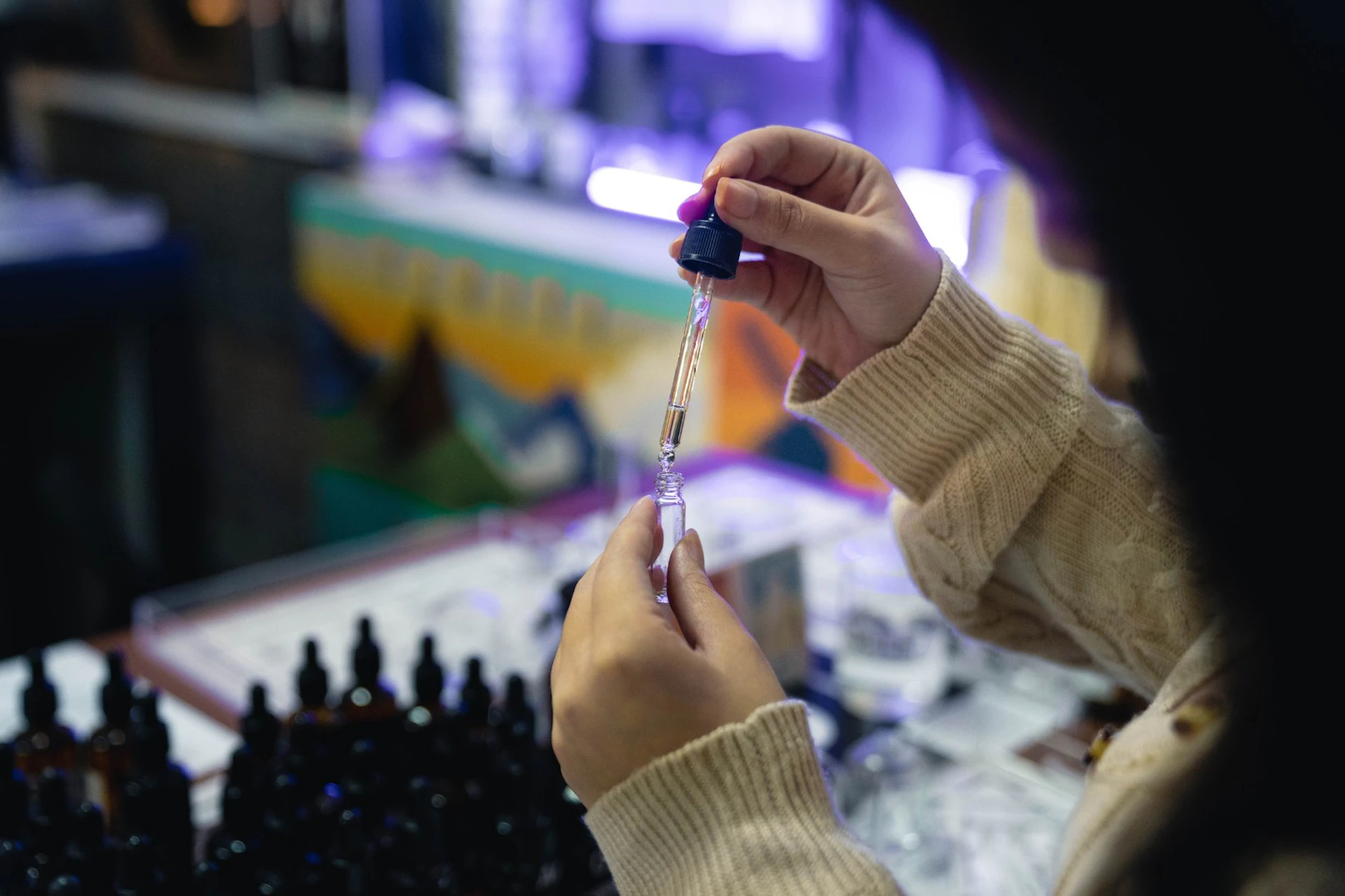 A person using a dropper to transfer liquid into a small vial, with rows of bottles on a table and a colorful background.