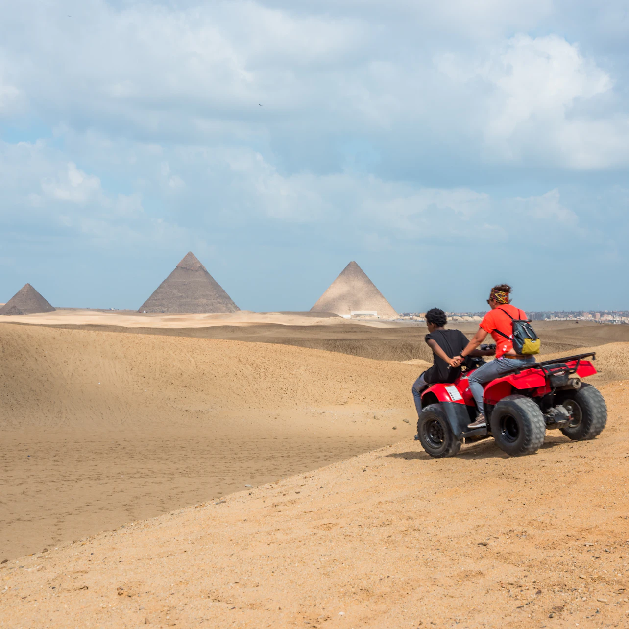 Two people ride an ATV in a sandy desert with pyramids in the background under a partly cloudy sky.
