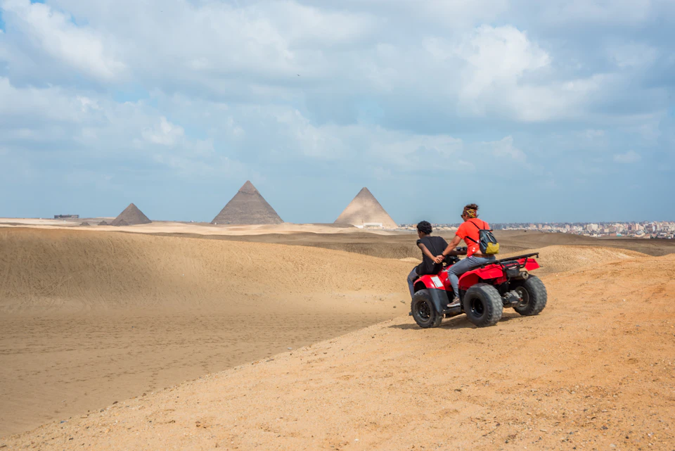 Two people ride an ATV in a sandy desert with pyramids in the background under a partly cloudy sky.