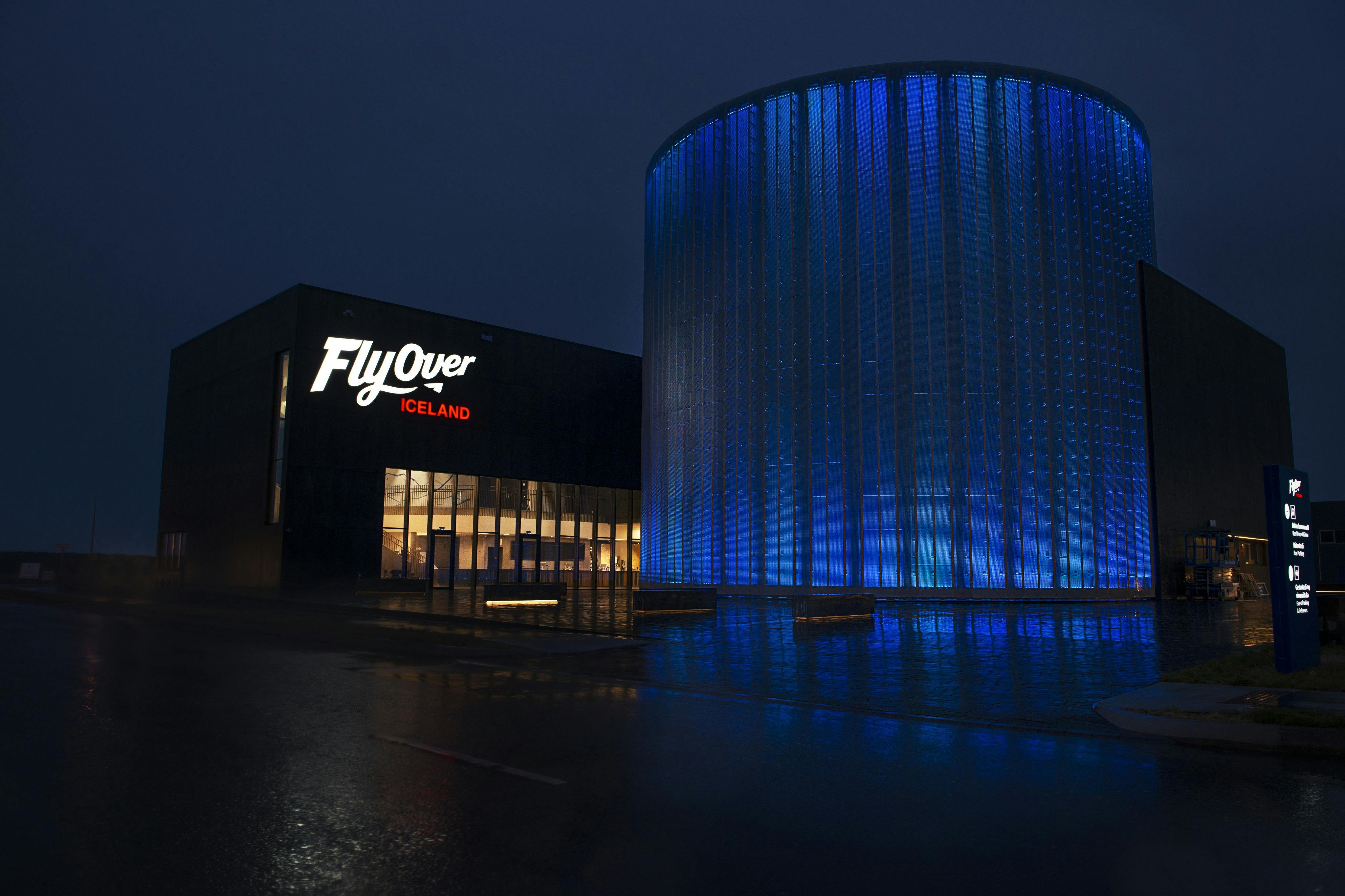 Beleuchtetes FlyOver-Island-Gebäude bei Nacht mit einer blau beleuchteten zylindrischen Struktur daneben.
