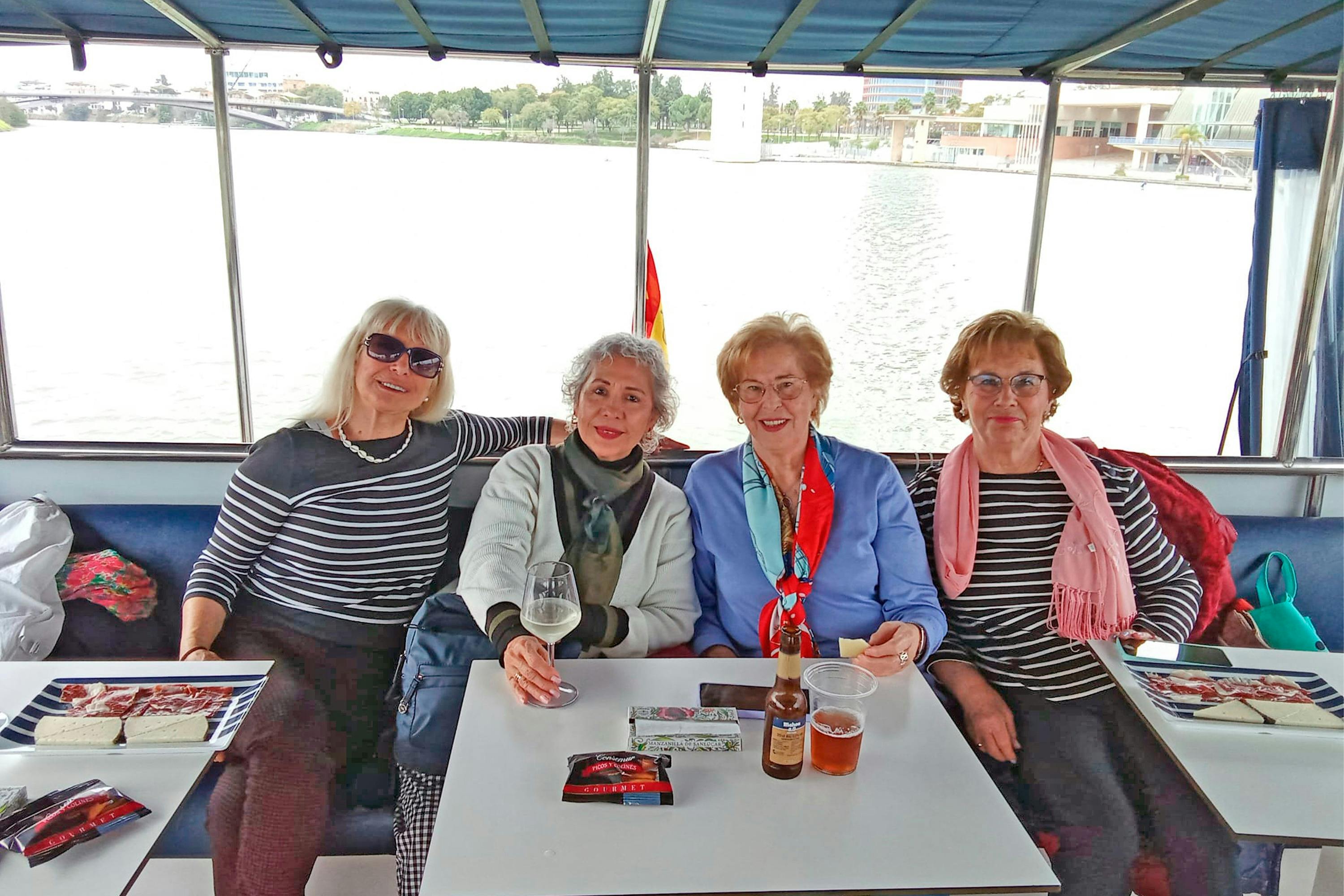 Drie vrouwen aan tafel op een boot, lachend, met drankjes en voorwerpen op tafel en water zichtbaar op de achtergrond.