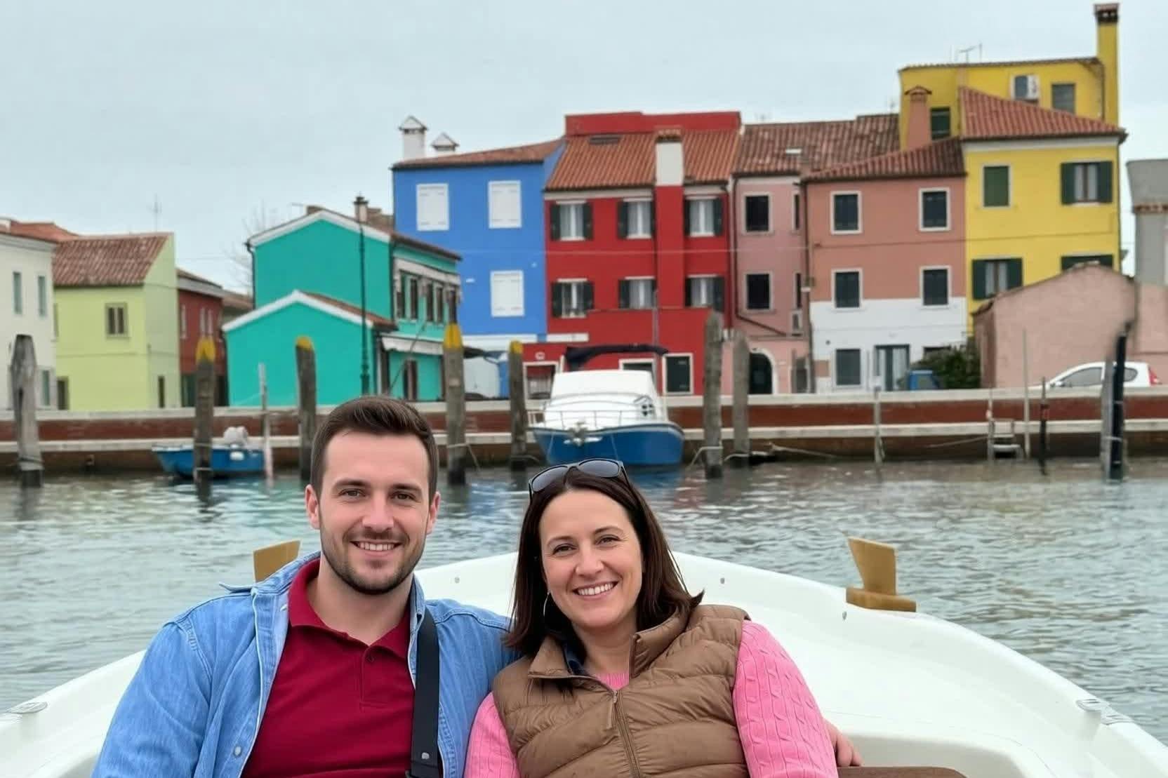 Un uomo e una donna sorridono seduti su una barca, con edifici colorati e un canale sullo sfondo.