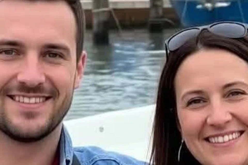 A man and a woman smiling while seated on a boat, with colorful buildings and a canal in the background.