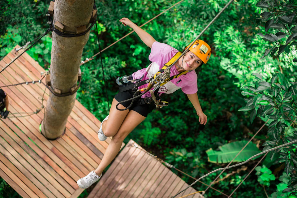 Person wearing a helmet and harness walking on a rope bridge in a lush green forest.