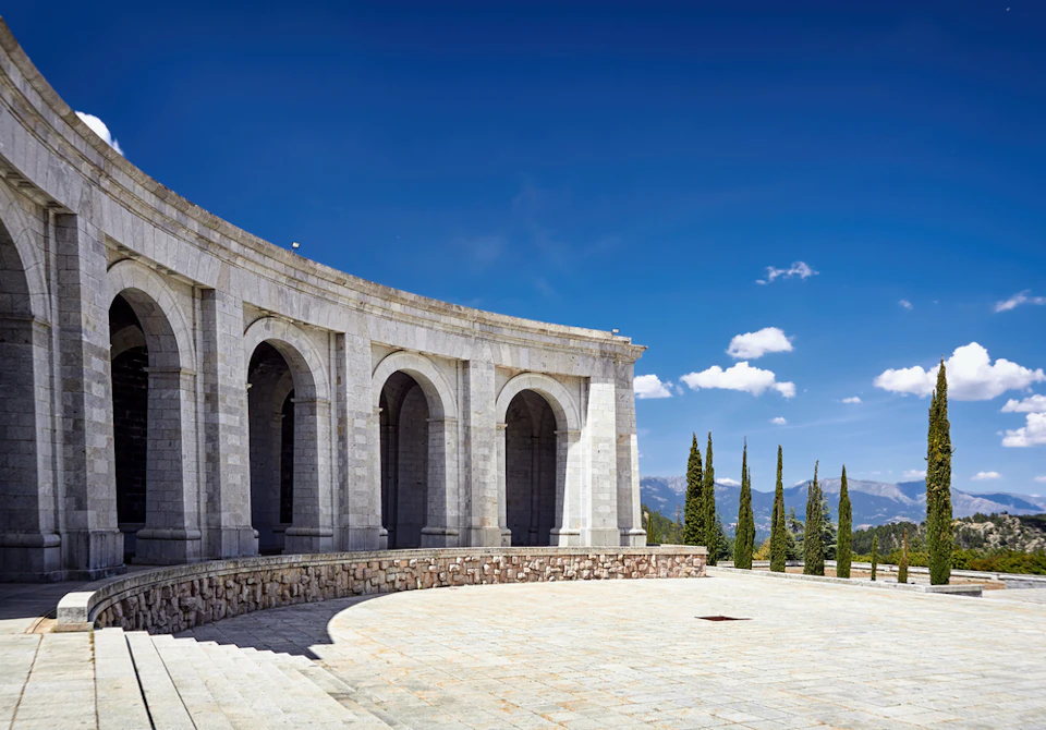 A stone building with large arches and a curved facade sits under a clear blue sky. Tall trees line the background.
