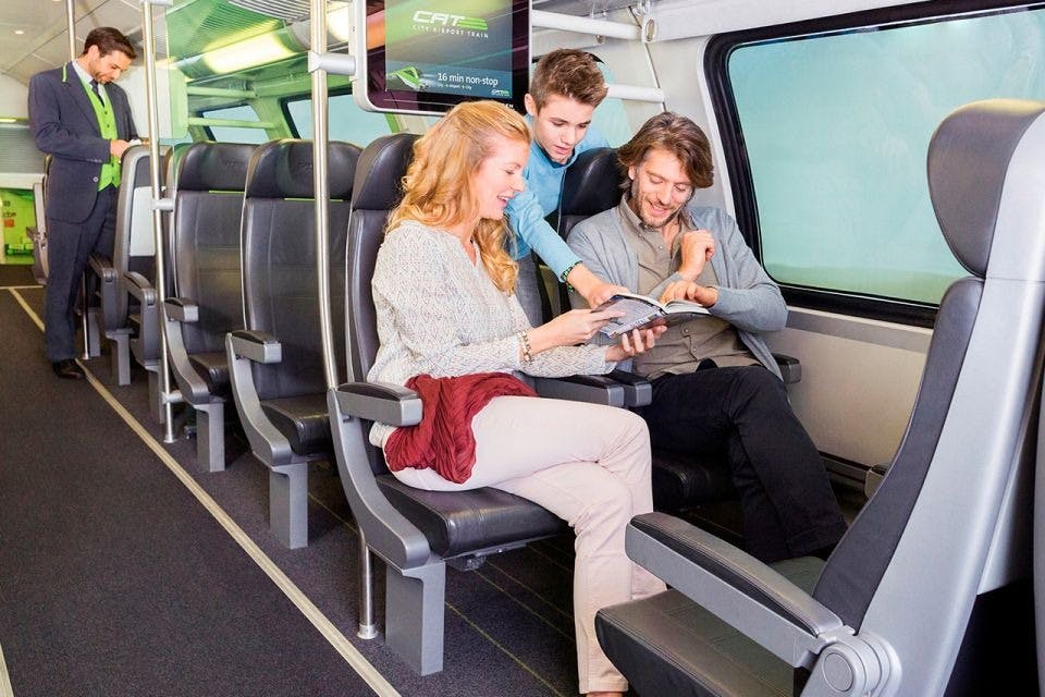 A family of three sits in a train, looking at a magazine; the child stands while the parents are seated next to each other.