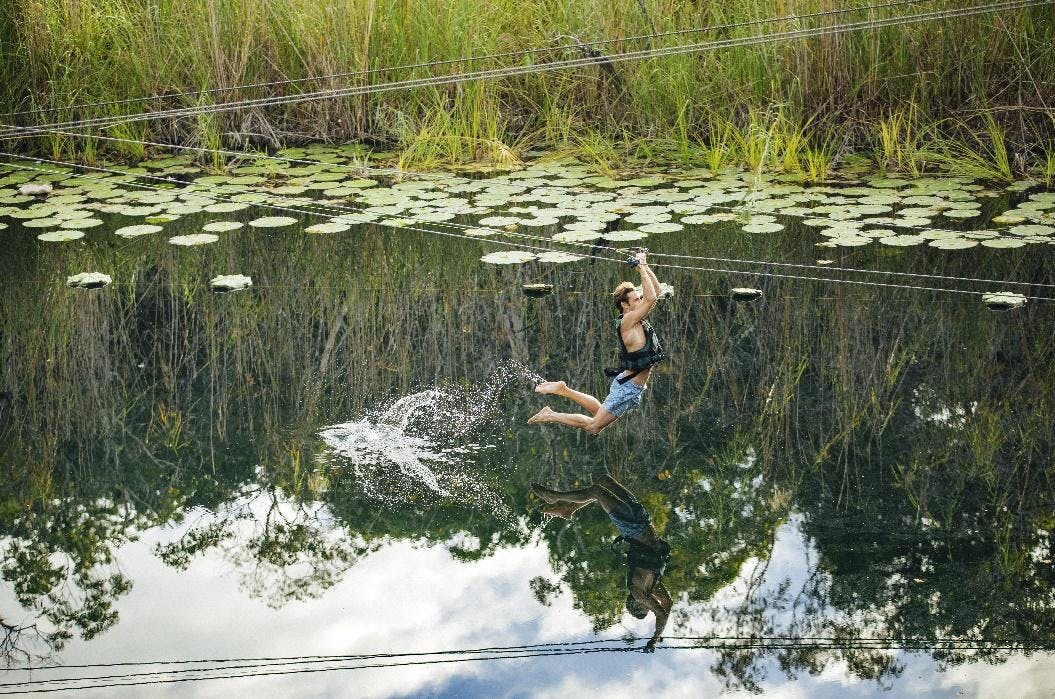 Osoba obložená zipem nad jezerem s liliovými polštářky a rákosím; stříkající voda pod nimi. Odražená obloha a stromy viditelné ve vodě.