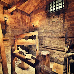 En svagt upplyst medeltida fängelsehåla med en stockad av trä, fackelbelysning, en stege och en figur med huva som håller en yxa.