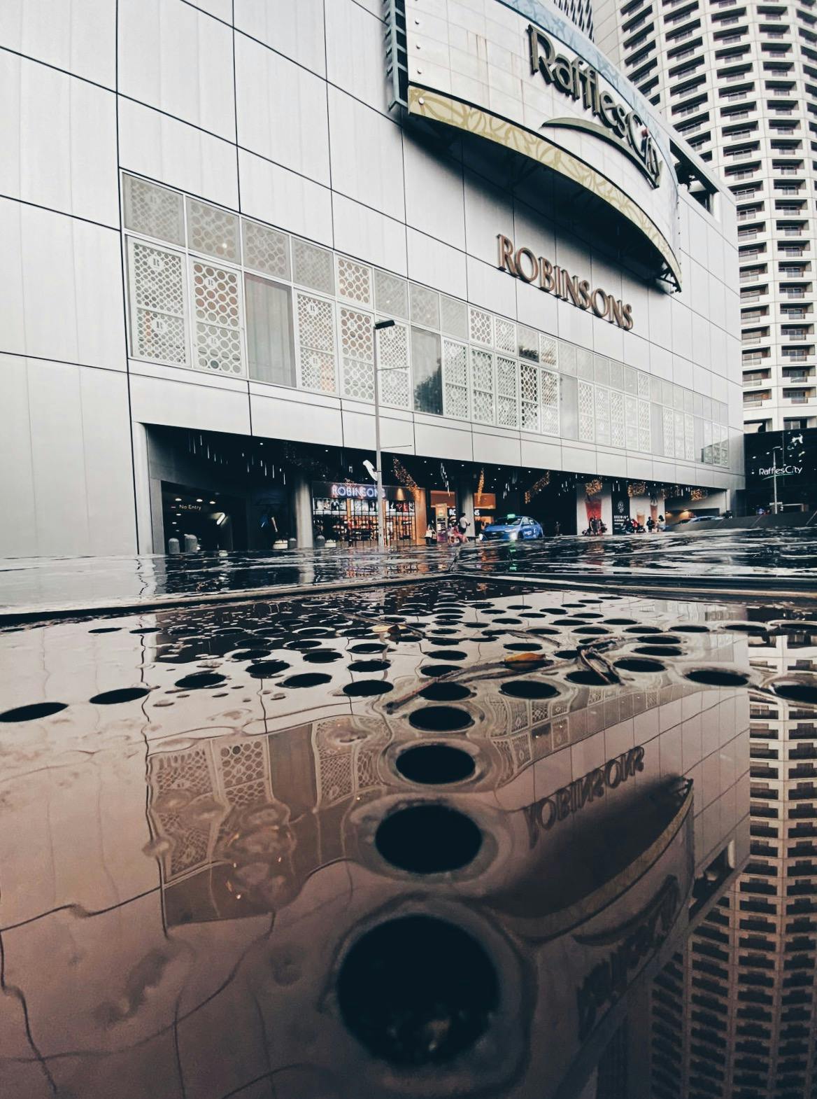 Una strada con una pozzanghera che riflette l'ingresso di un edificio con l'insegna "ROBINSONS". Vicino all'ingresso sono visibili persone e automobili.