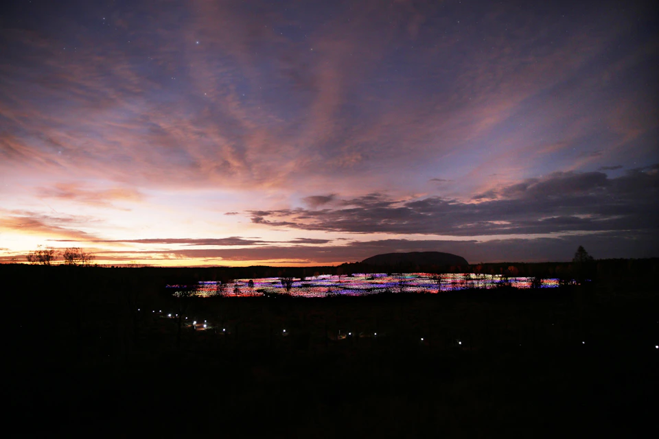 A landscape at dusk with a horizon of colorful lights and a cloudy sky, featuring a large silhouette of a distant hill.