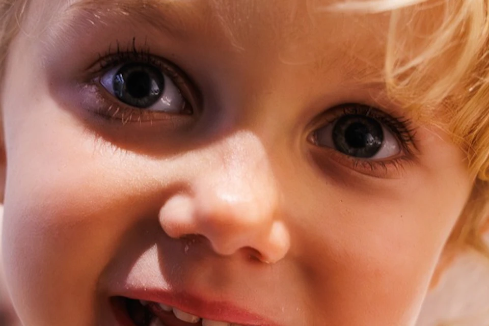 A child with curly blonde hair wearing a yellow sweater is smiling and holding a small object, with a blurry indoor background.