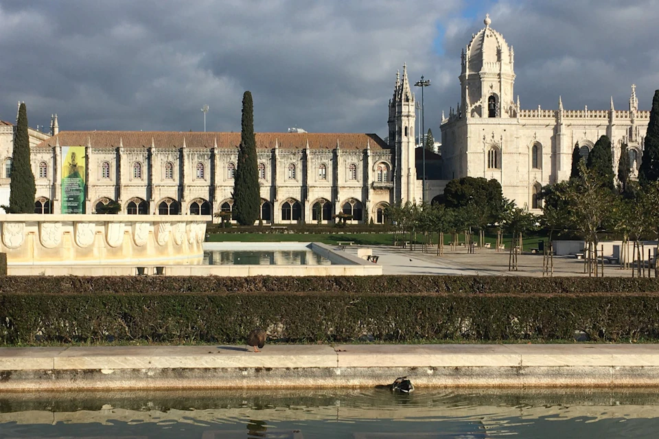 Historic monastery with Gothic architecture, a large tree, a reflecting pool with fountains, and ducks in the foreground. Cloudy sky overhead.