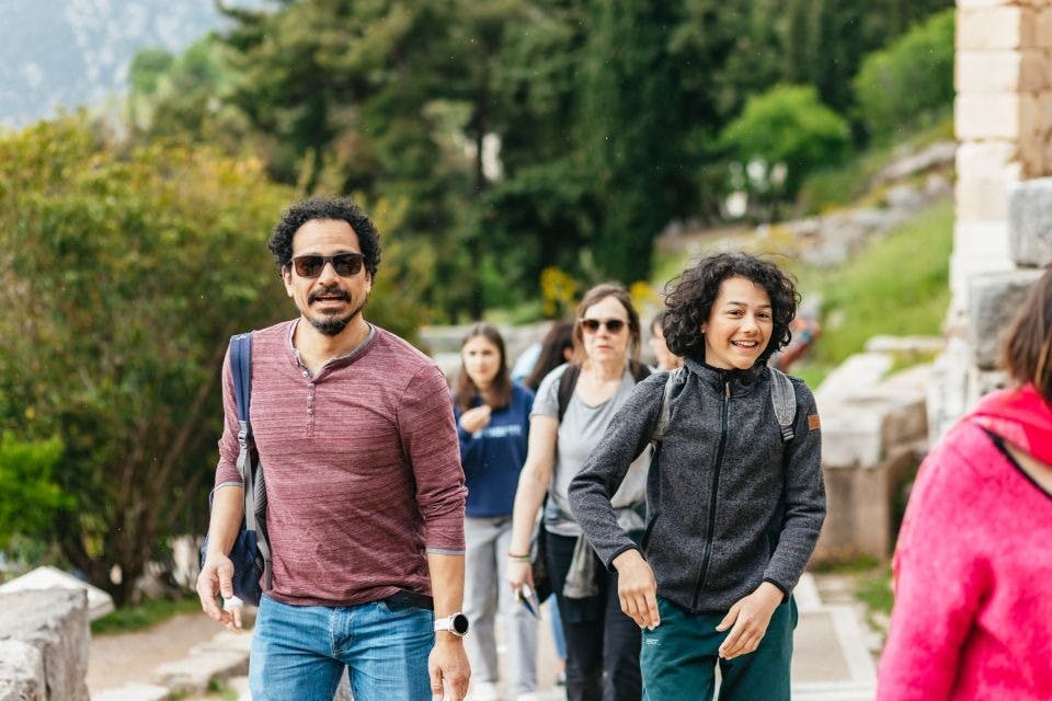 녹음으로 둘러싸인 길에서 야외를 걷고 있는 한 무리의 사람들. 빨간 셔츠를 입은 남자와 웃는 젊은이가 앞에 있습니다.