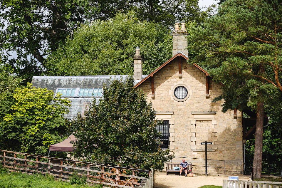 A man sits on a bench outside a stone building surrounded by trees and greenery on a sunny day.