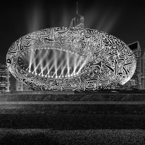 Illuminated ring-shaped structure with intricate script, skyscrapers, and light beams in the background at night.