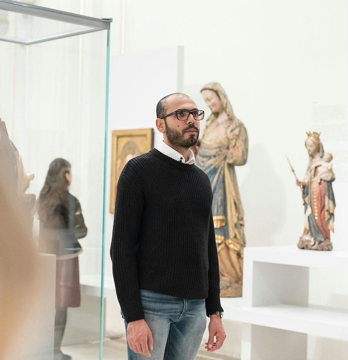 Muž v černém svetru a brýlích stojí v muzeu umění poblíž náboženských soch a zarámovaných uměleckých děl. Jiný se dívá na exponáty.