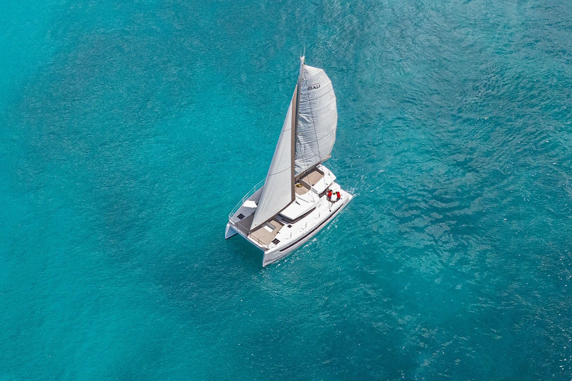 A white sailboat with one sail glides on clear, turquoise water, viewed from above.