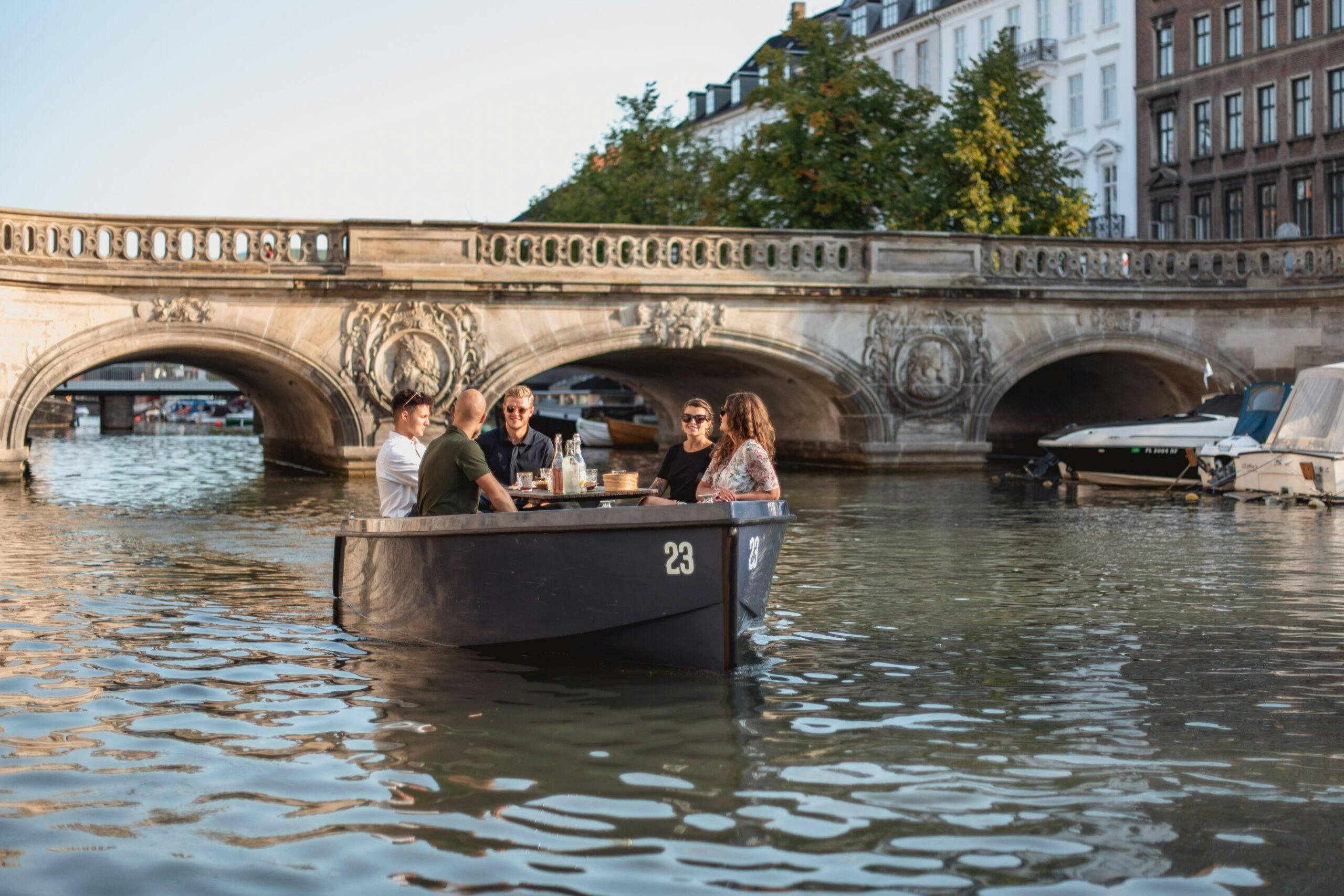 Five people sitting in a small boat on a canal, conversing and drinking. An ornate stone bridge is in the background.