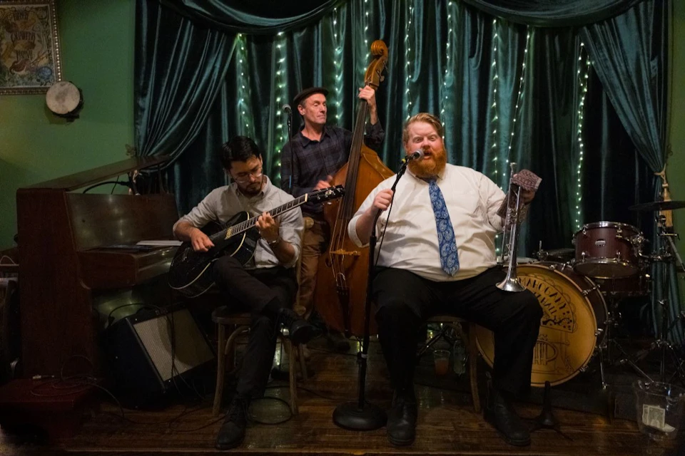 Three musicians perform on stage: one plays guitar, one upright bass, and one sings into a microphone while holding a trumpet.