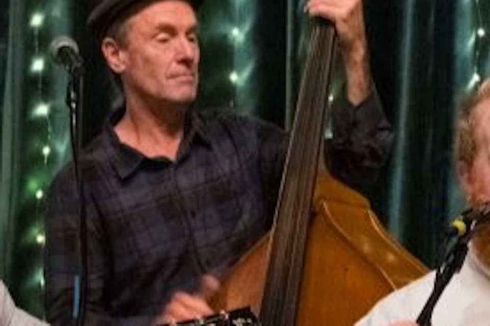 Three musicians perform on stage: one plays guitar, one upright bass, and one sings into a microphone while holding a trumpet.