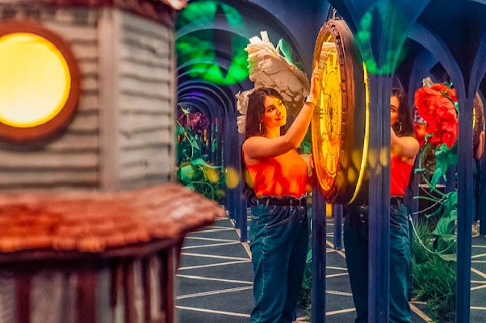 A woman in a vibrant setting interacts with a glowing circular object, surrounded by reflective surfaces and colorful decor.