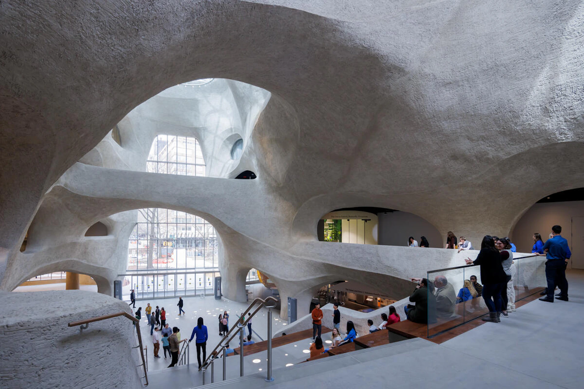 Spacious modern atrium with curved concrete architecture, glass walls, staircases, and people walking and mingling.