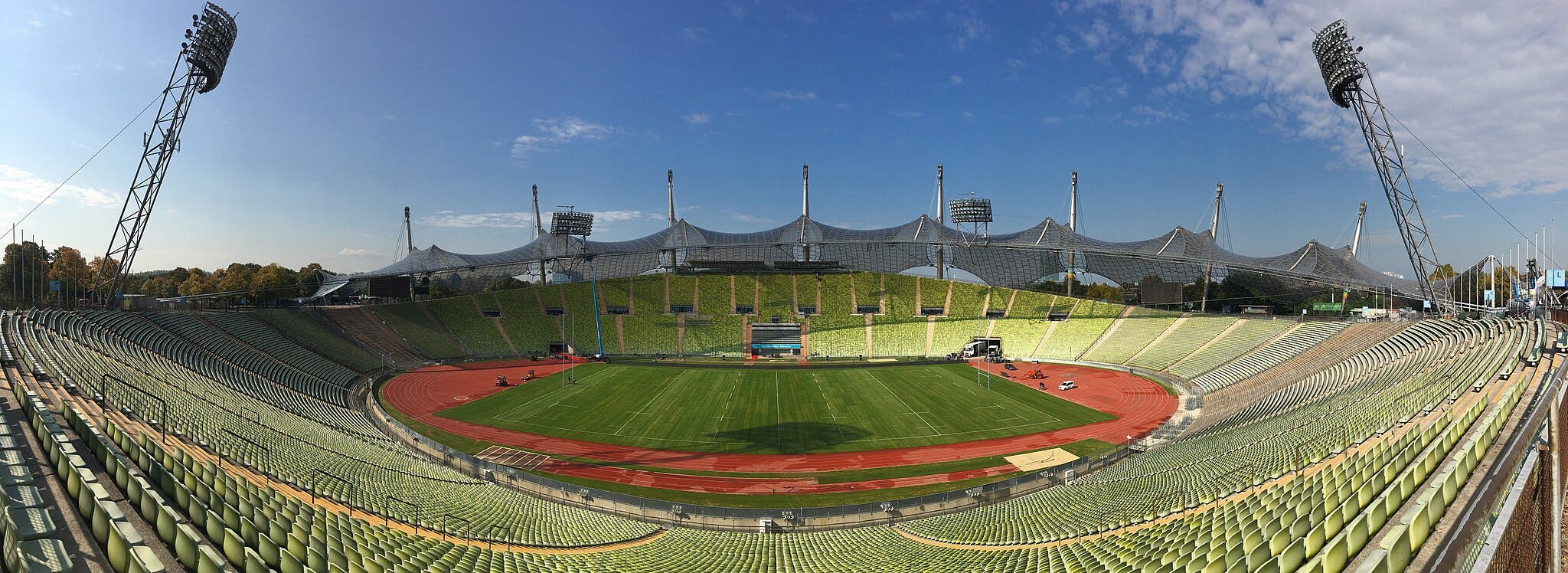 Panoramic view of an empty stadium with green seats, a grass field, and distinctive architectural roof supports under a blue sky.