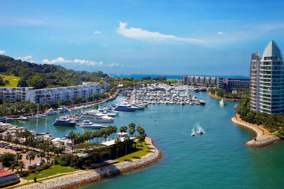 A marina with numerous docked boats, surrounded by buildings, greenery, and calm waters under a clear blue sky.