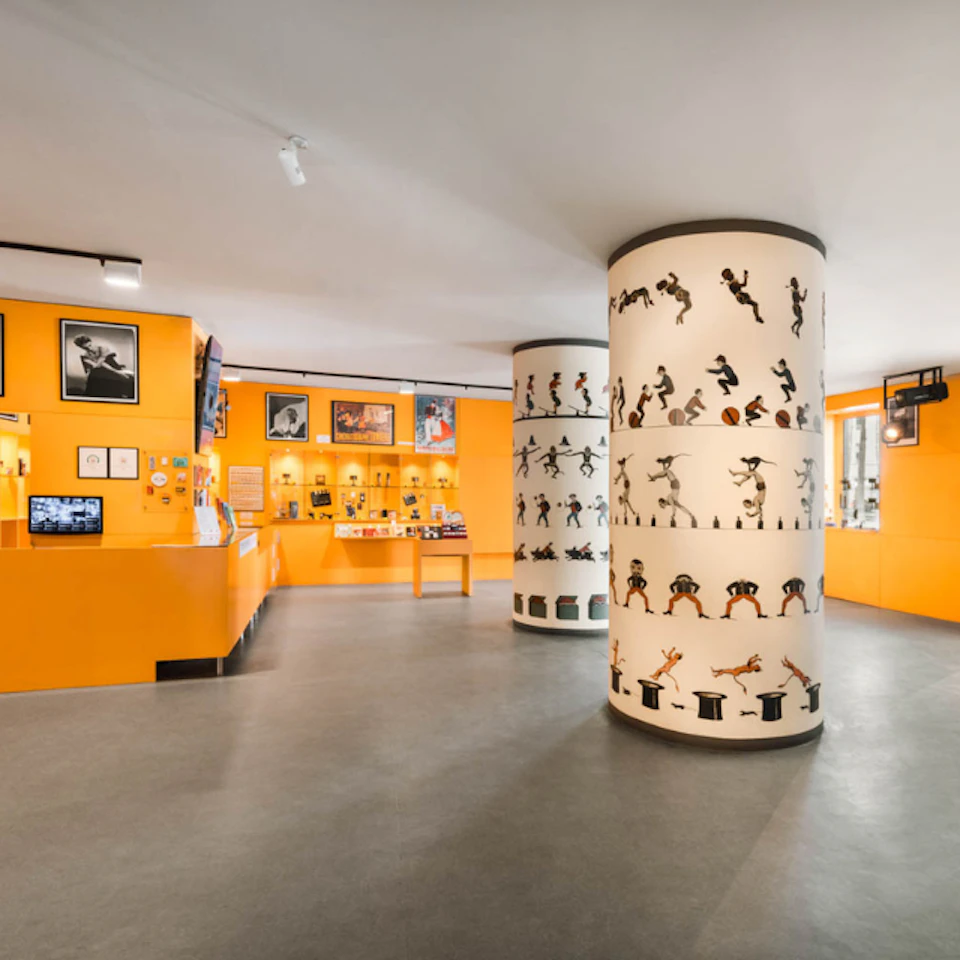 A brightly lit room with orange walls features display cases, framed photos, and columns wrapped in dancing character animations.