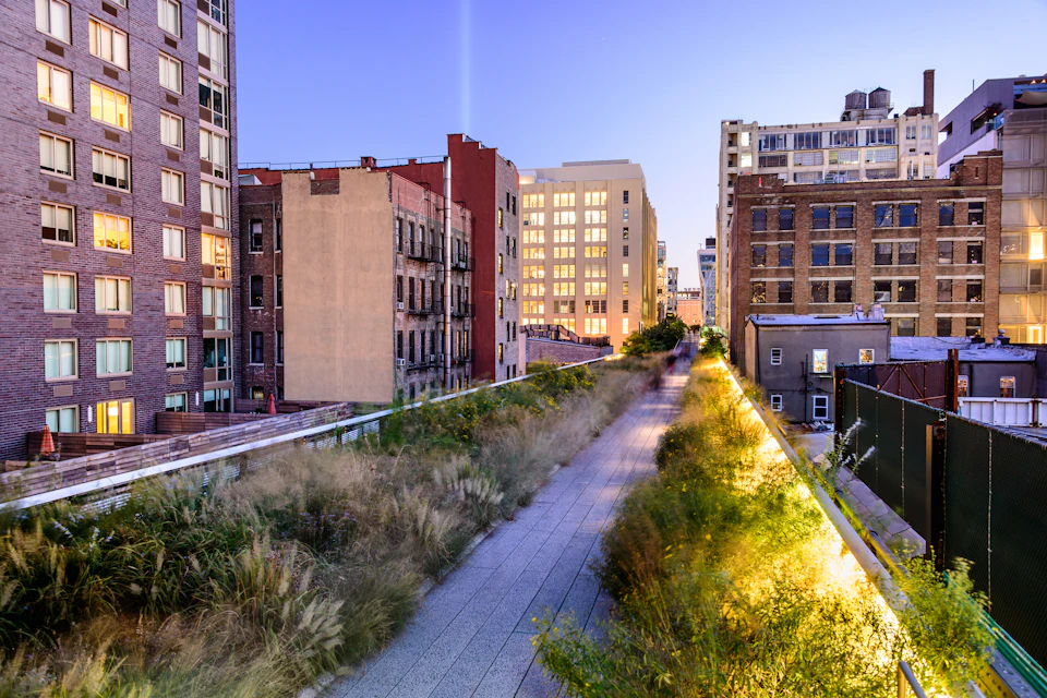 Elevated urban park with plants and a walkway surrounded by buildings at dusk, with soft lighting illuminating the pathway.