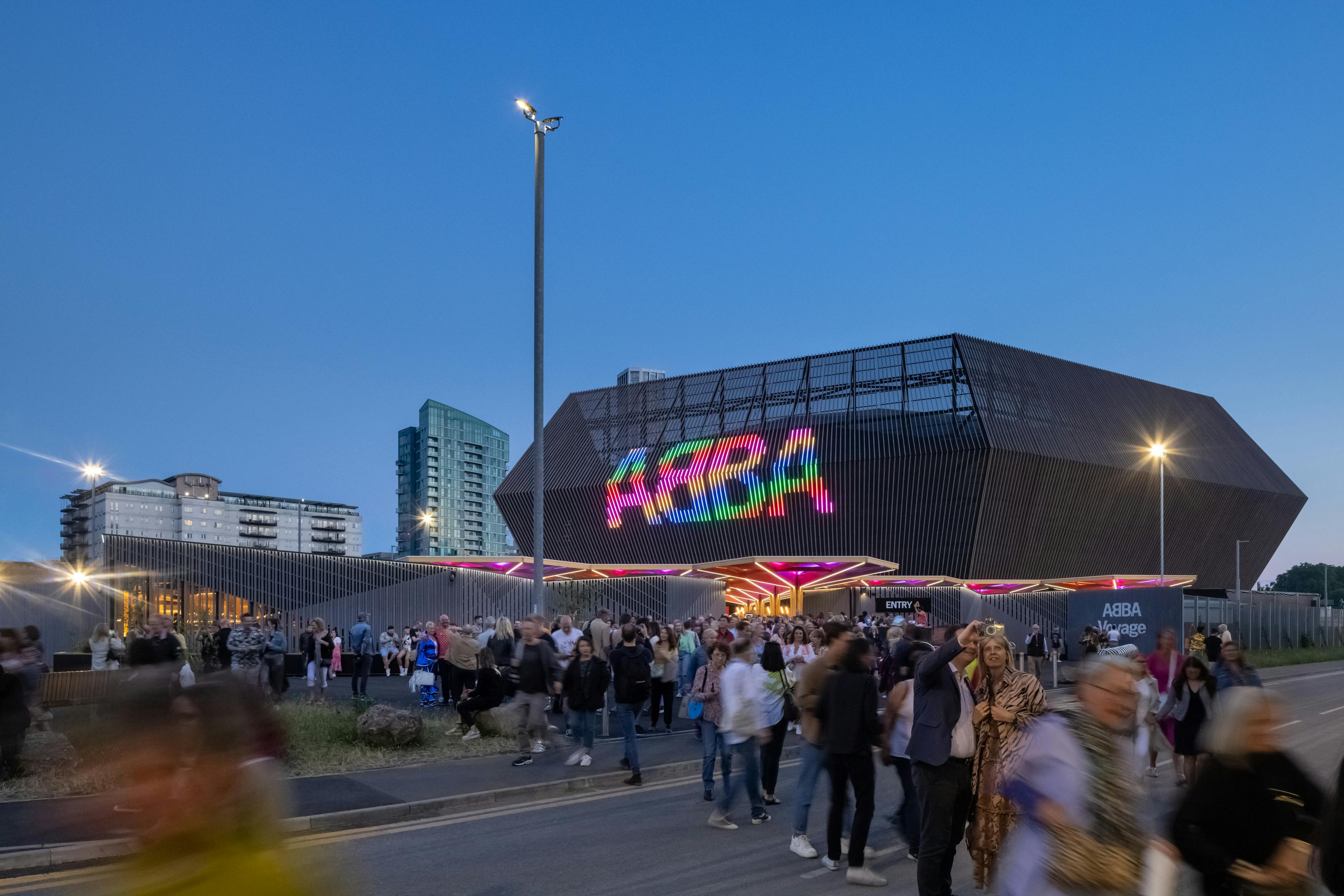 People gathering outside a modern, illuminated venue with a colorful “ABBA” sign at dusk, with tall buildings in the background.