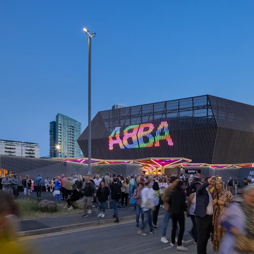 Människor som samlas utanför en modern, upplyst plats med en färgstark "ABBA" -skylt i skymningen, med höga byggnader i bakgrunden.