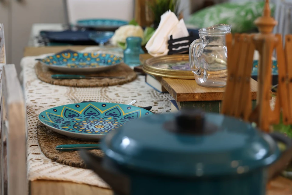 Table set with colorful plates, a glass pitcher, folded napkins, and a blue pot lid in the foreground.