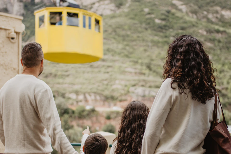 A family looks at a yellow cable car against a green, hilly backdrop.