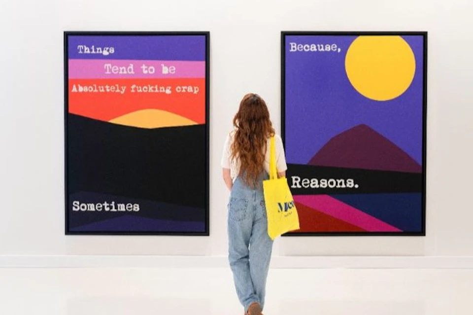 A person with long hair and a yellow tote bag looks at two colorful abstract art pieces with text on them in a gallery.