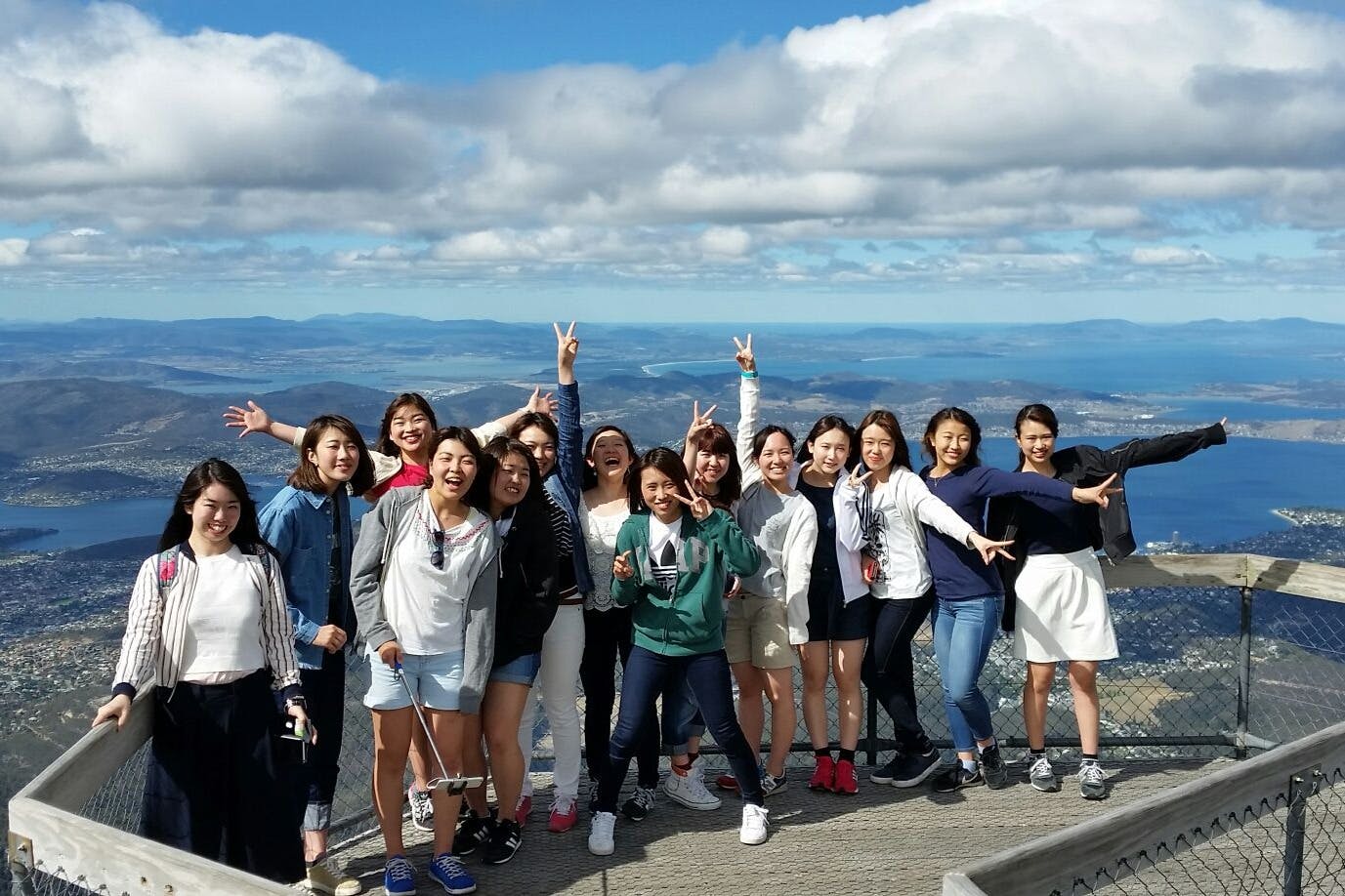 A group of people smiling and posing on a mountain viewpoint with a scenic ocean and islands in the background on a partly cloudy day.