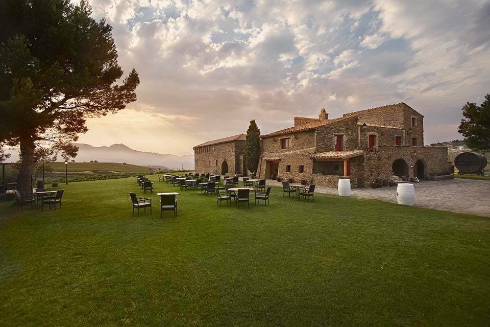 Rustic stone building with red shutters, surrounded by outdoor tables and chairs on a green lawn, under a cloudy sky at sunset.