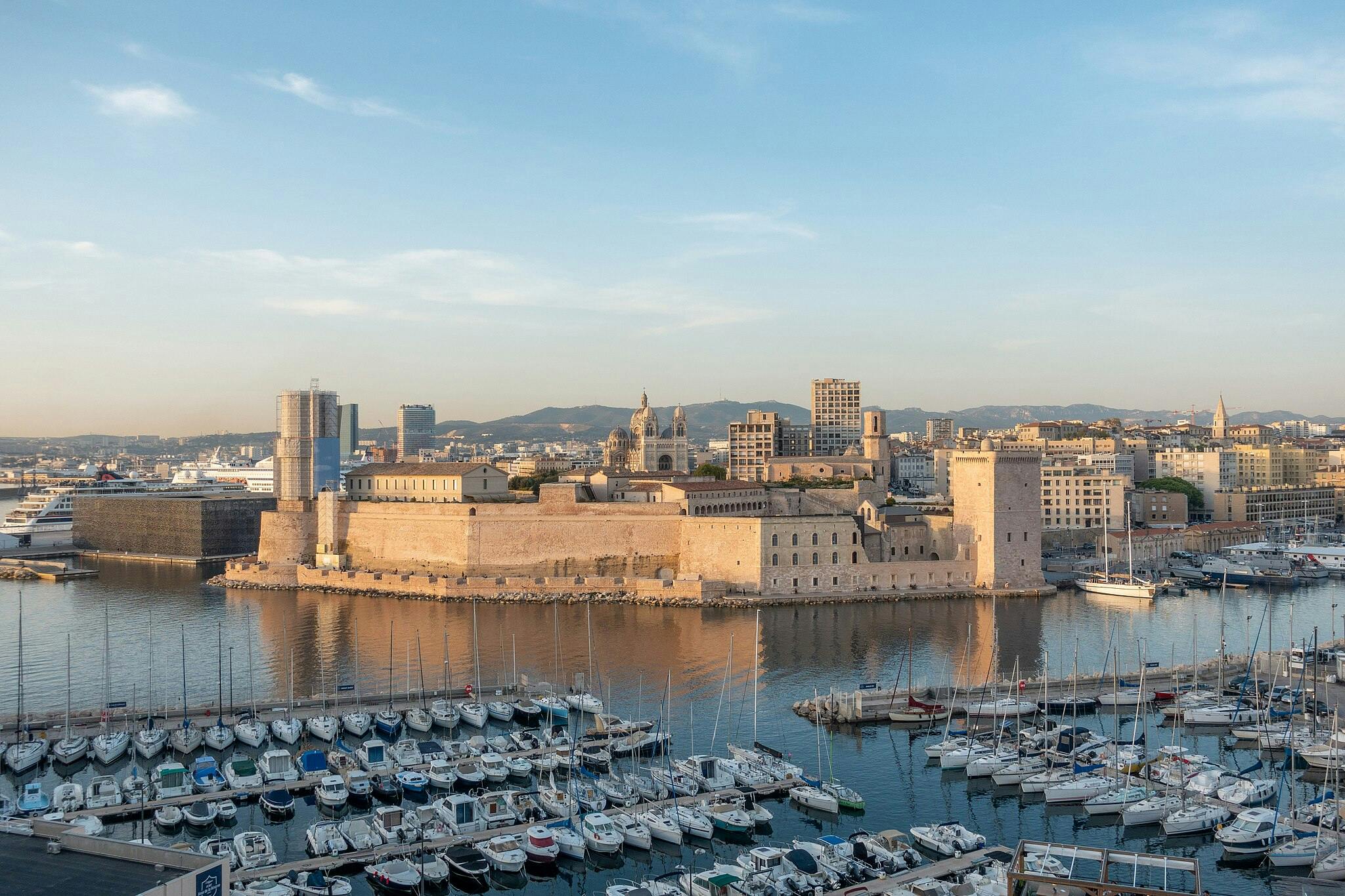 A coastal cityscape with a large historic fortress, numerous docked boats, and surrounding modern buildings under a clear sky.