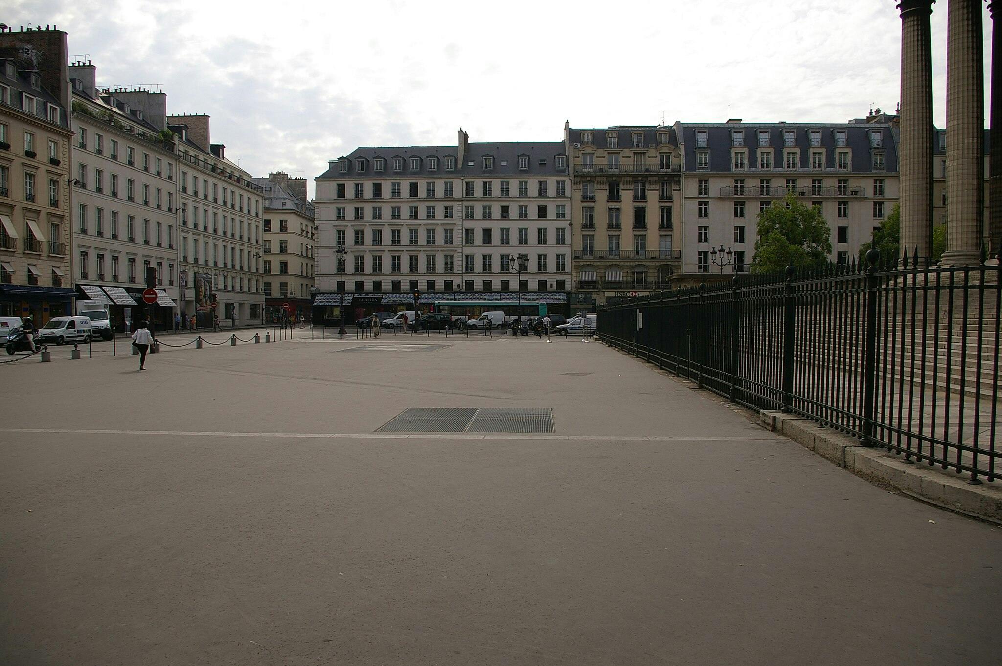 복층 건물과 검은색 철제 울타리로 둘러싸인 몇 사람이 걸어다니는 대부분 텅 빈 도시 광장입니다.