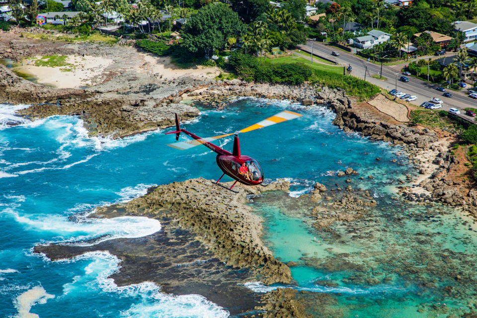 A red helicopter flies over a rocky coastline and turquoise ocean, near a road and green trees.
