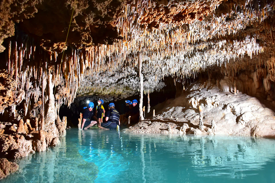 Several people wearing helmets with headlamps explore a cave with stalactites and stalagmites, standing in shallow, clear water.