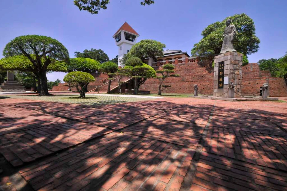 A historic site with a red brick courtyard, a white watchtower, green trees, and a statue under a clear blue sky.