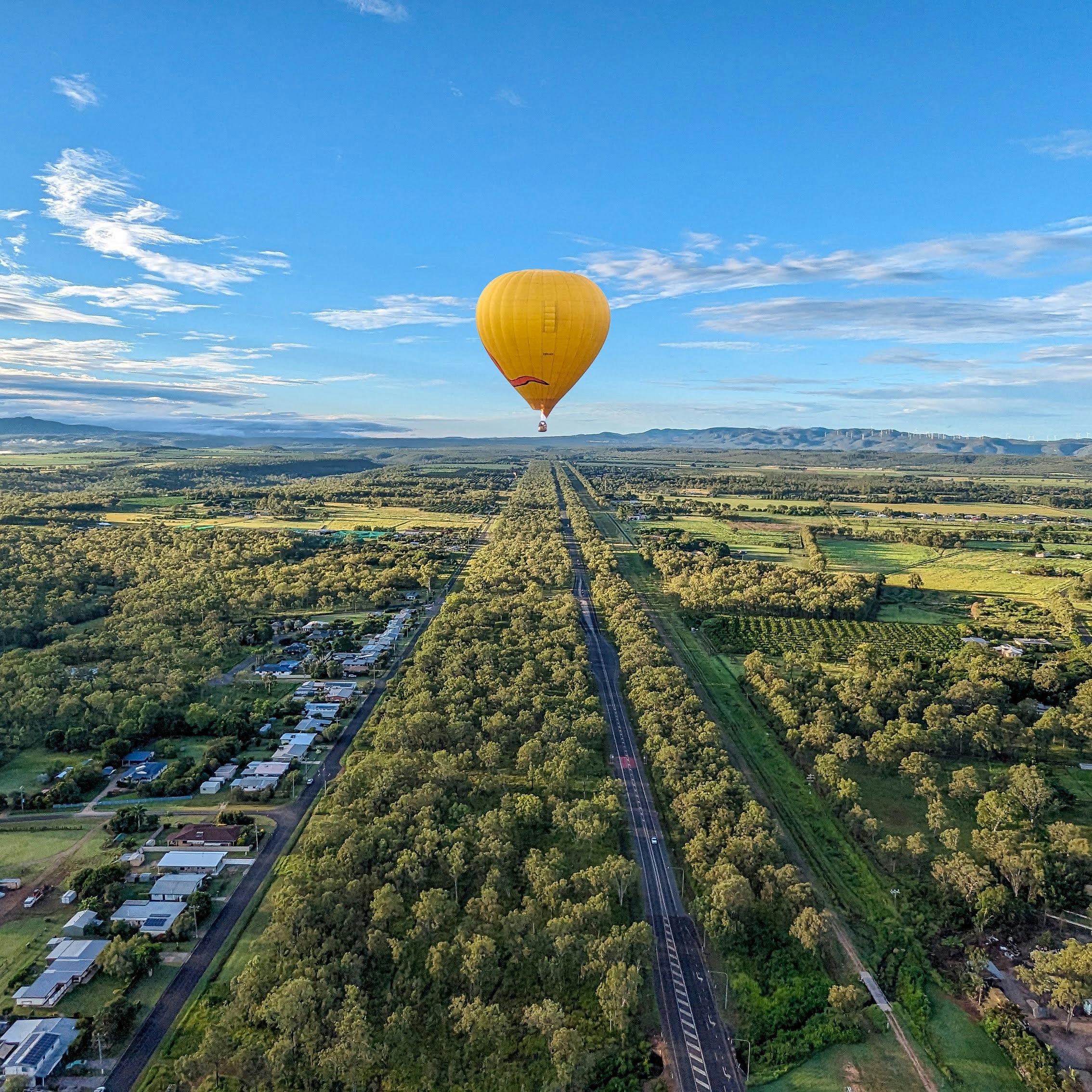 Two hot air balloons, one yellow and one orange, float above a field at sunrise with a forest in the background.