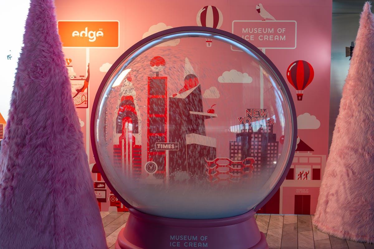 Velká sněhová koule se stylizovaným panoramatem města s růžovými tóny, vystavená v místnosti s barevnými stěnami a dekoracemi.