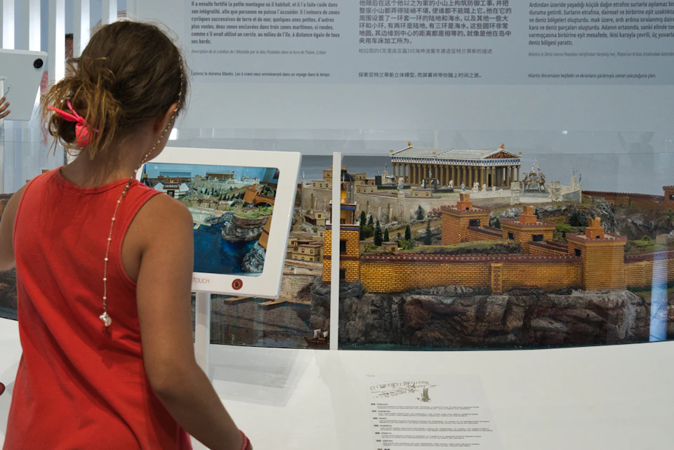 A person in a red sleeveless shirt interacts with a touchscreen display showcasing a detailed historical model.