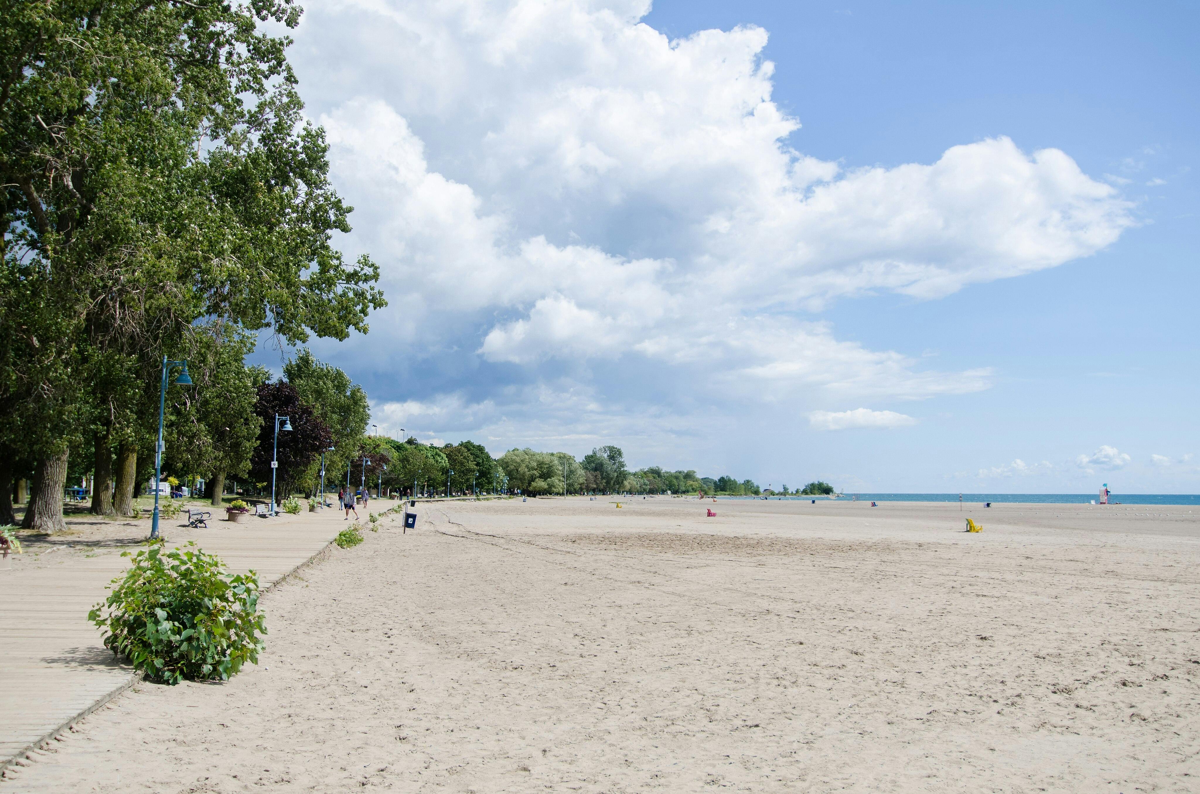 Empty sandy beach with a boardwalk lined with trees and benches on the left, and ocean waves under a partly cloudy sky.