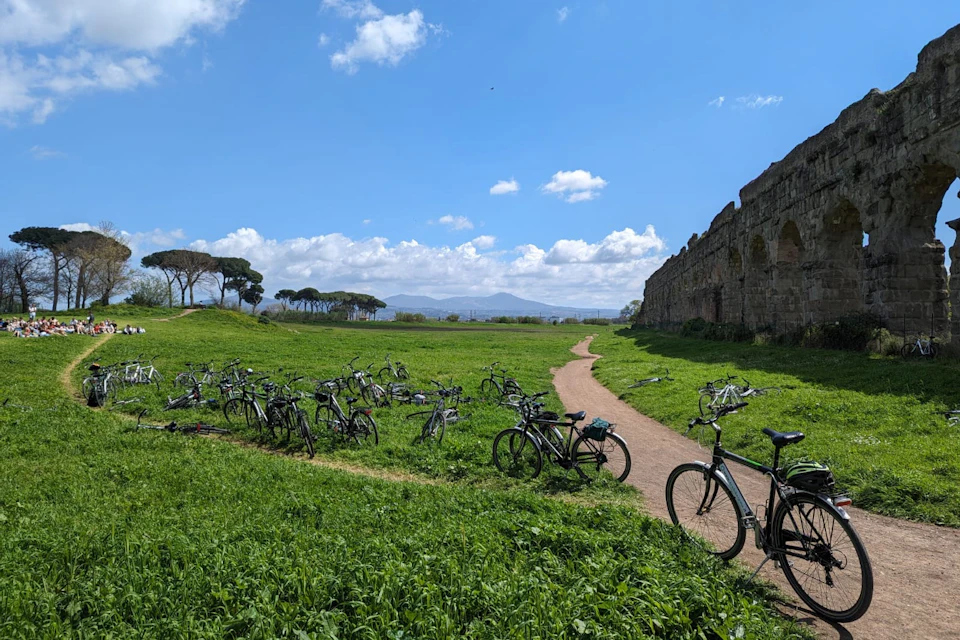 A scenic grassy area with a dirt path, numerous parked bicycles, trees, and ruins under a blue sky with clouds.
