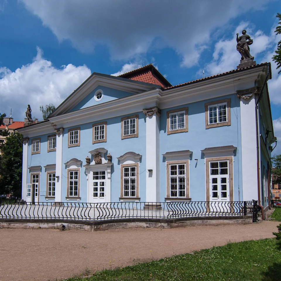 Light blue, two-story building with white trim, multiple windows, and statues on the roof, under a partly cloudy sky. Pathway and railing in front.