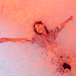 En person som ler och ligger i en stor grop fylld med genomskinliga rosa plastkulor.