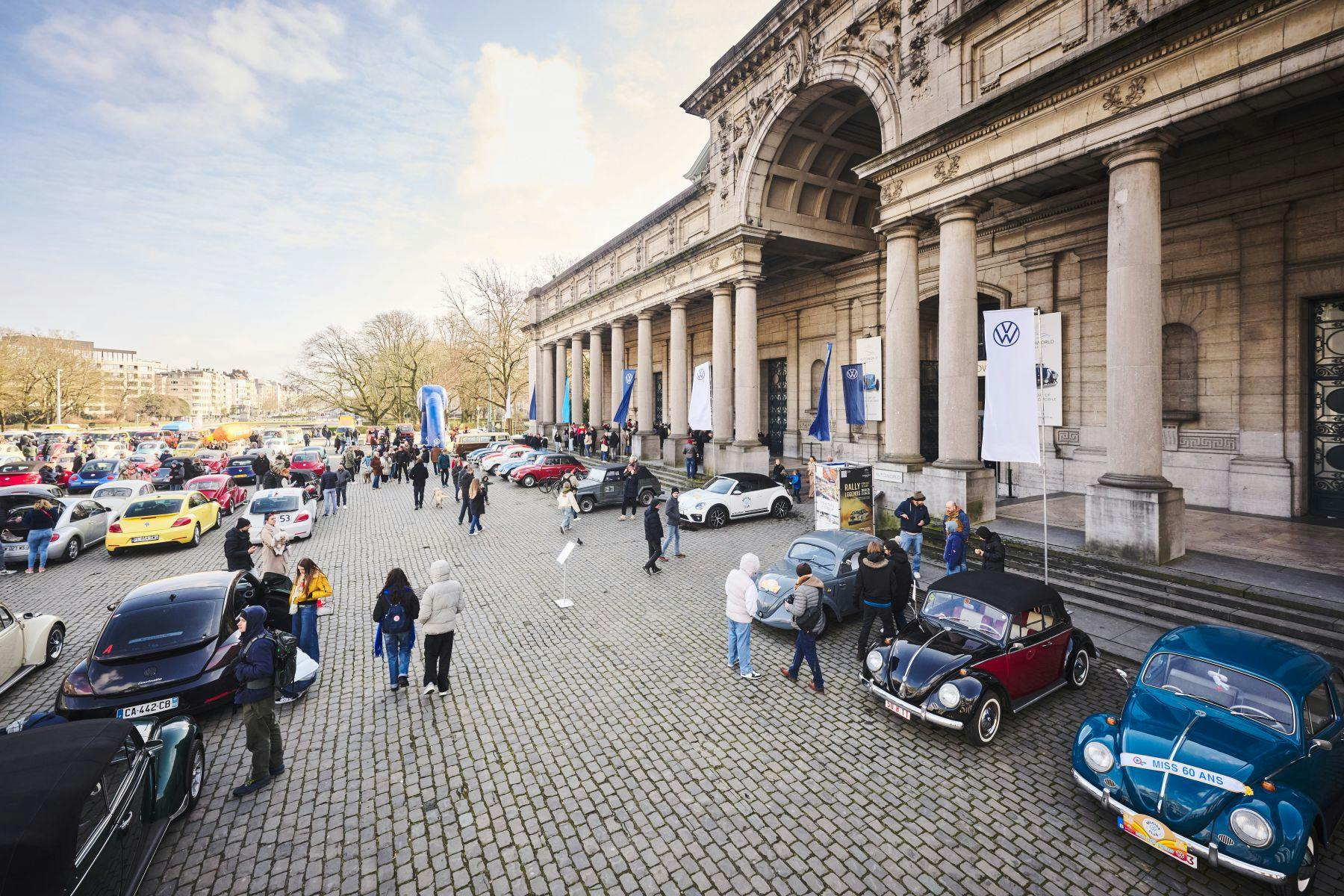 Des personnes parcourent des voitures anciennes devant un grand bâtiment historique avec des colonnes et des bannières Volkswagen.